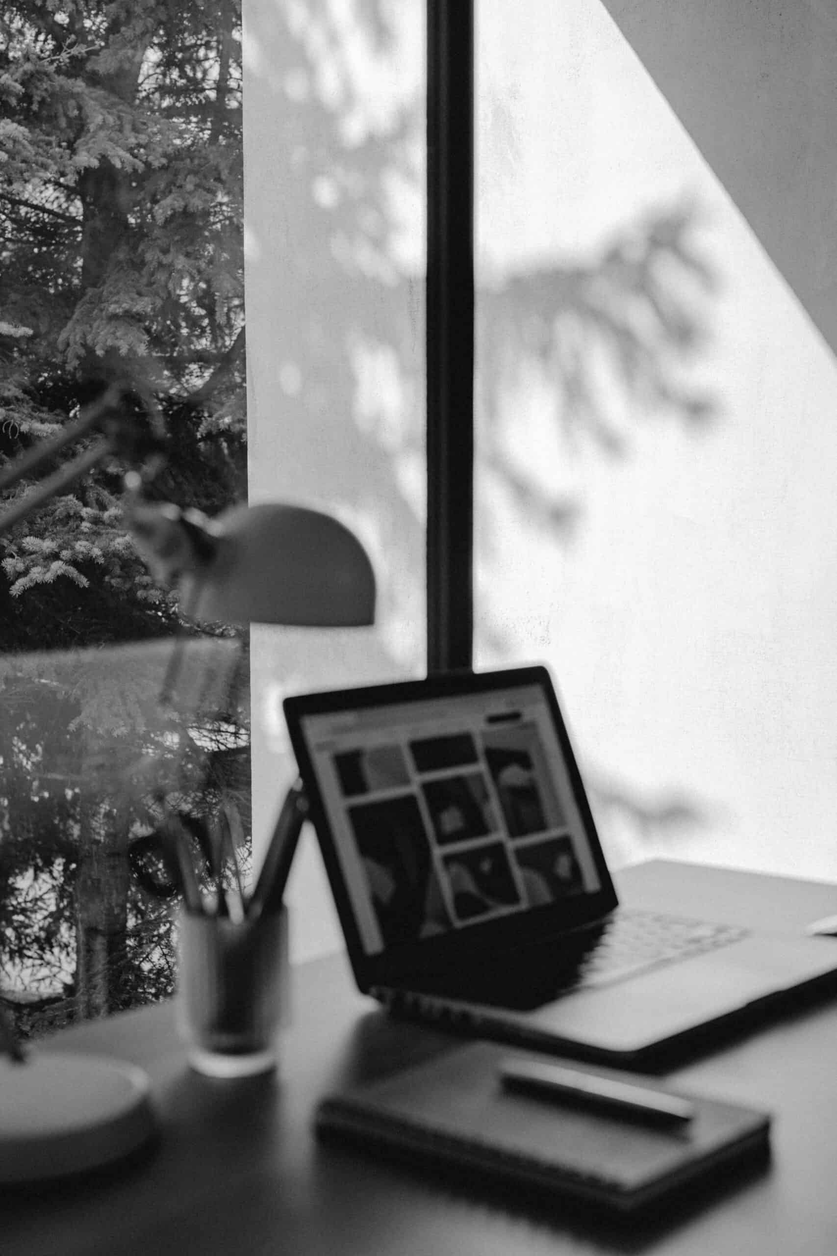 A black-and-white photo of a desk by a window, featuring a laptop, a lamp, a cup with pens, and a notebook. Sunlight and tree shadows are cast on the wall, creating a calm, minimalist workspace.