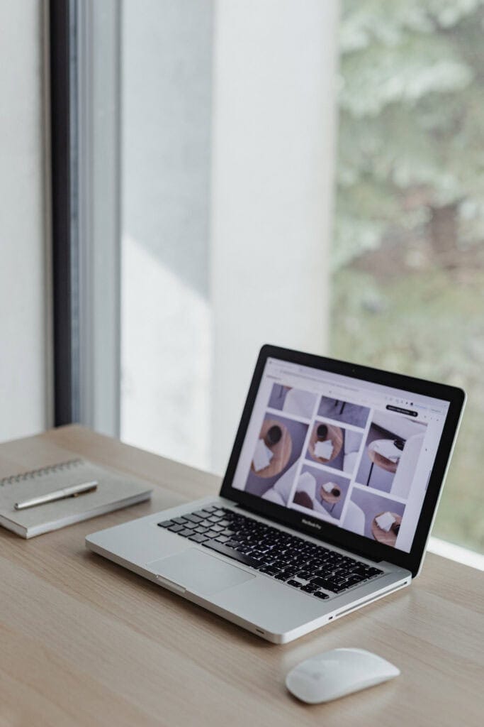 A laptop displaying a grid of photos sits on a light wooden desk next to a notebook, pen, and computer mouse. The desk is by a large window with a blurred view of greenery outside.