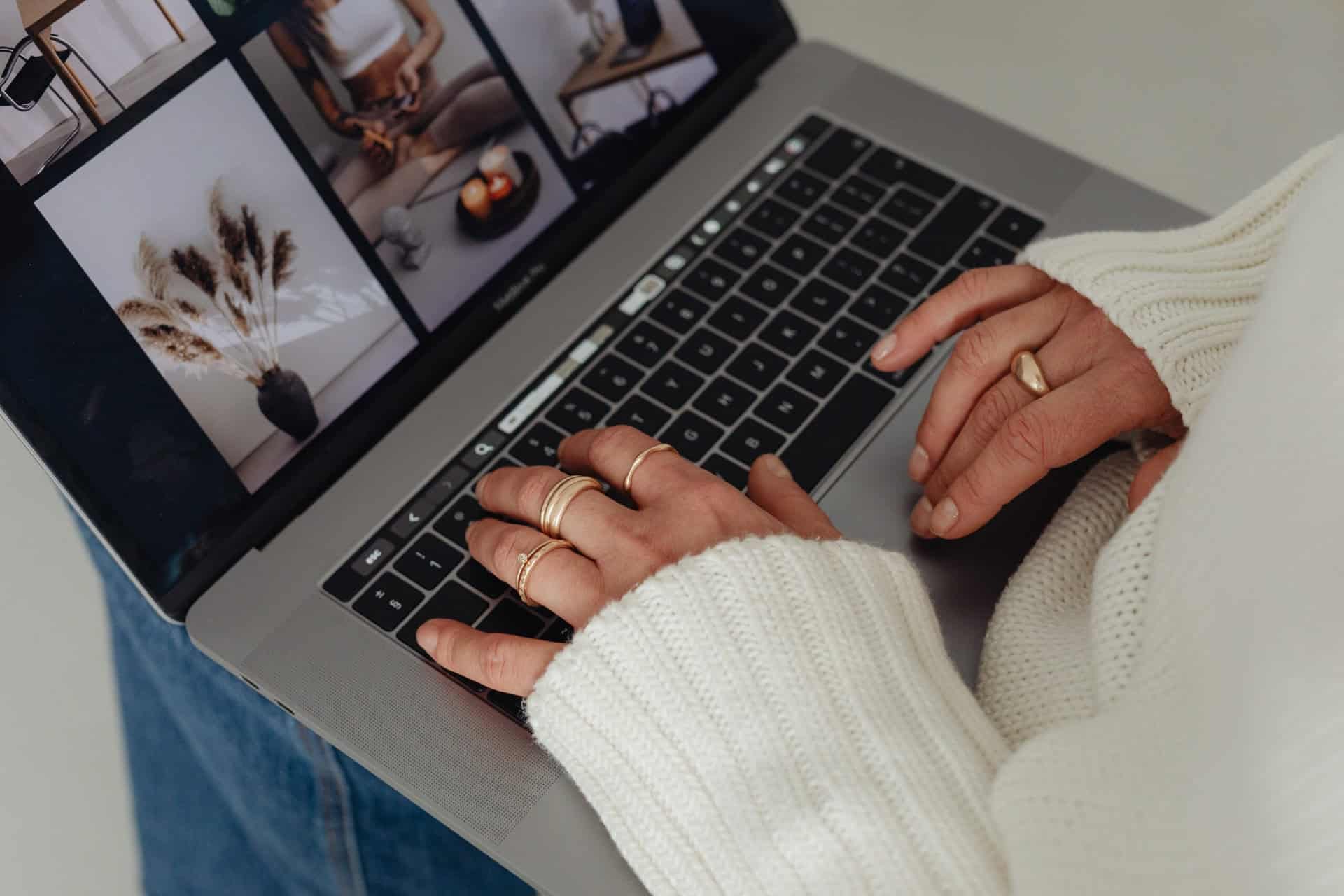 A person wearing a white sweater and several gold rings types on a laptop keyboard. The laptop screen displays several aesthetic photos, including home decor and plants.